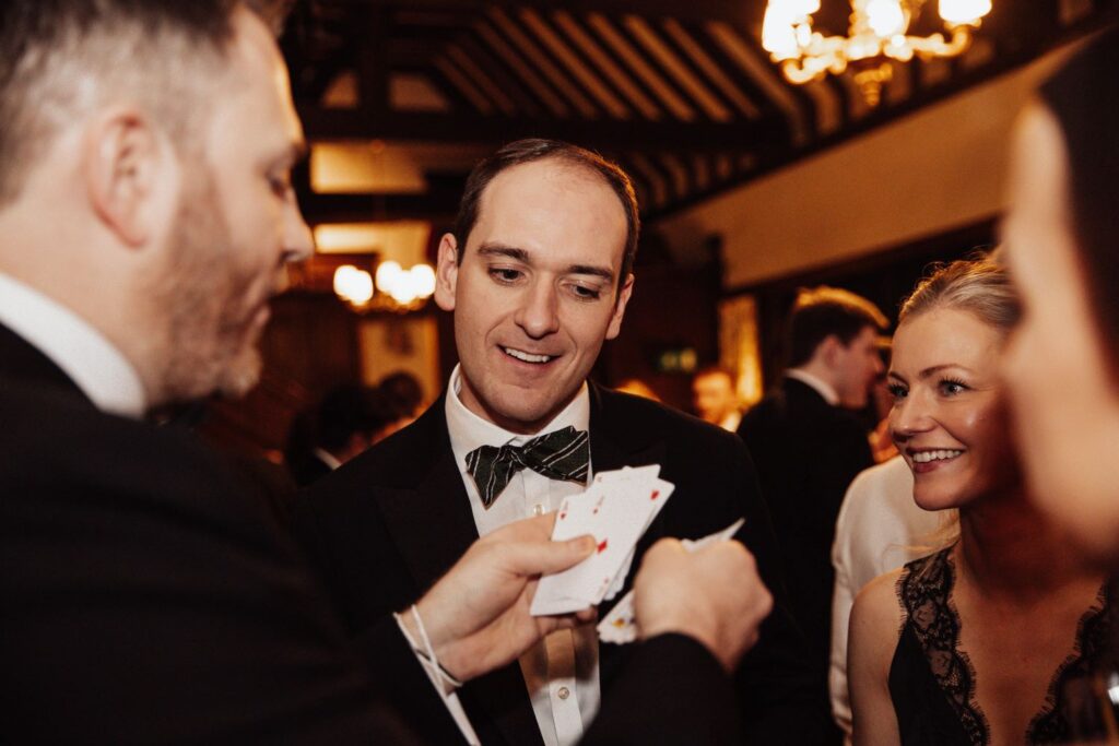 Magician handling a pack of cards while guests smile and watch his hands closely. Black Christmas party event.